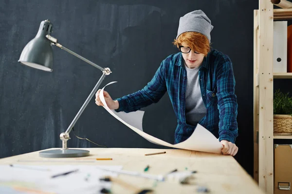 ビーニー帽子とオフィスで黒板背景に青写真と一緒に作業中机に立ってメガネを身に着けている若い創造的な赤髪の男の肖像 — ストック写真