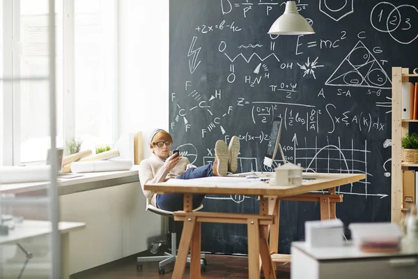ビーニー帽子とメガネの科学的な数式をチョークで書かれた黒板に対してスマート フォンを使用して テーブルの脚と椅子で休んでいる身に着けている赤髪青年の肖像画 — ストック写真