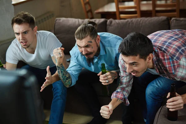 在电视上观看体育比赛的三情绪化成年男子的画像和兴奋的欢呼 而坐在沙发上的客厅里的啤酒 — 图库照片