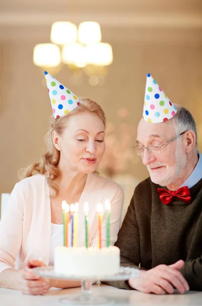 幸せな成熟した男とケーキのキャンドルを燃焼を見て誕生日キャップで彼の妻 — ストック写真