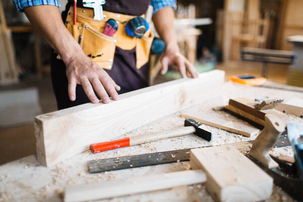 Close-up shot of male hands examining surface of wooden board, hammers, planes and saw lying on table
