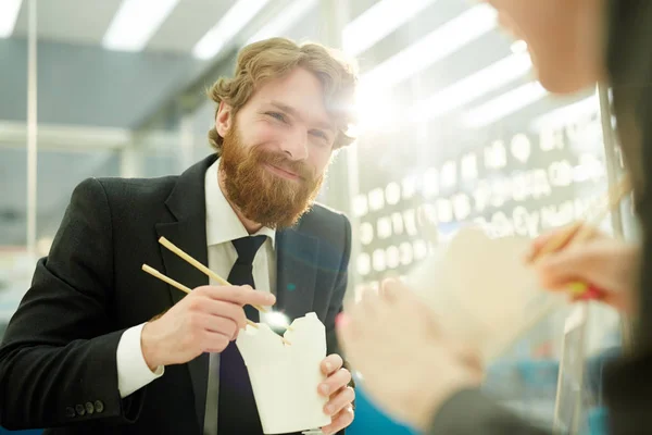 Portrait of modern bearded businessman smiling happily while eating Chinese food in office sharing lunch with colleague