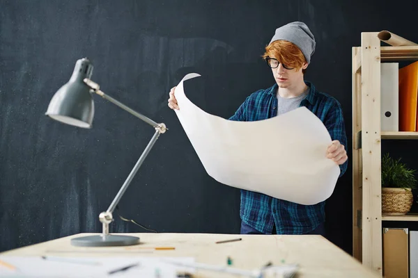 ビーニー帽子とオフィスで黒板背景に青写真と一緒に作業中机に立ってメガネを身に着けている若い創造的な赤い髪のエンジニアの肖像画 — ストック写真