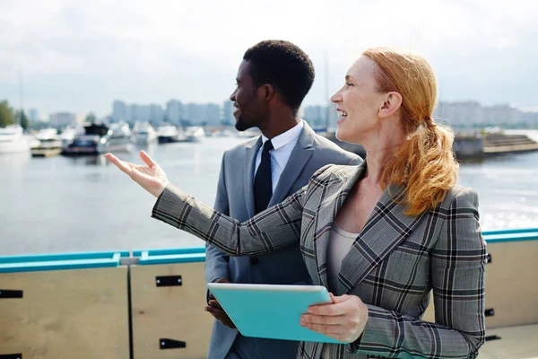 Profile view of attractive middle-aged businesswoman pointing at something while standing on upper deck with African American colleague and discussing joint project, she holding digital tablet in hand