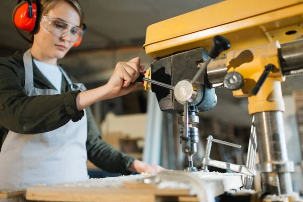 Attractive young carpenter in apron looking at wooden work piece with concentration while working with drill press, waist-up portrait