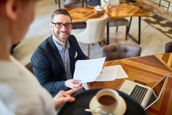 Friendly bearded financial manager distracted from paperwork and smiling to waitress while working at modern cafe, she holding tray with coffee cup