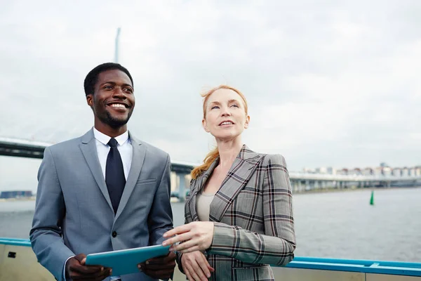 Two confident colleagues in formalwear looking away while standing on upper deck of ship and brainstorming on start-up project, African American manager holding digital tablet in hands