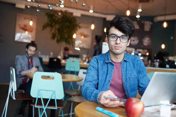 カフェのテーブルにノート パソコンとスマート フォンを操作しながらカメラ目線のメガネとジーンズのジャケットを着てハンサムな若い男の肖像 — ストック写真
