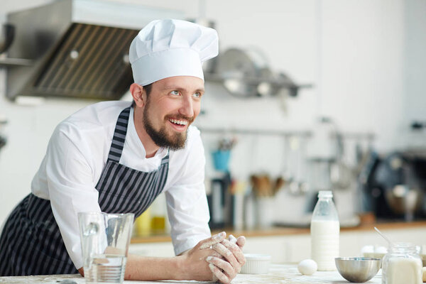 Cheerful baker or pastry-chef leaning at his workplace with ingredients for dough