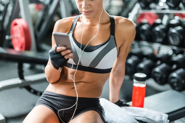 不辨认的适合的被晒黑的妇女的中间部分射击穿着运动服坐在健身房与智能手机 锻炼后休息 — 图库照片
