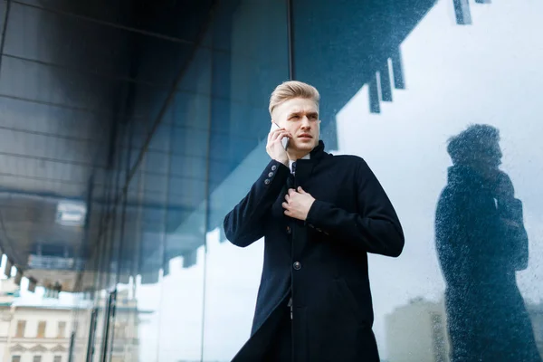 Portrait of concentrated white collar worker having difficult phone conversation while standing against glass outer wall of office building, low angle view