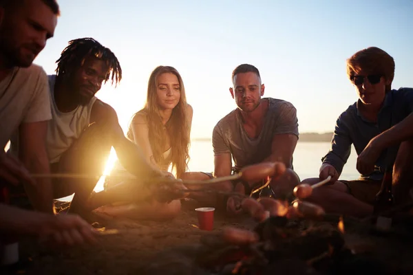 Multi-ethnic friends making snack on campfire while relaxing by water