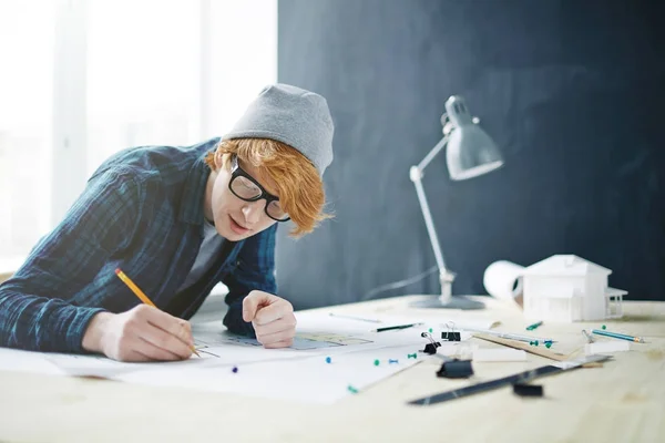 年轻的红头发设计师的肖像戴着眼镜和帽子在办公室工作 靠在办公桌上的灯和房子模型 绘制平面图 — 图库照片
