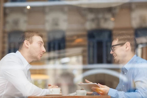 两个人在咖啡馆见面的侧面图坐在桌对面和讨论工作 从玻璃窗口后面射击 — 图库照片