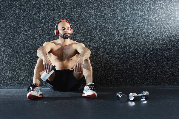 强壮肌肉男子的肖像坐在地板上饮水和听音乐的大耳机 而休息后高强度锻炼在健身房 — 图库照片