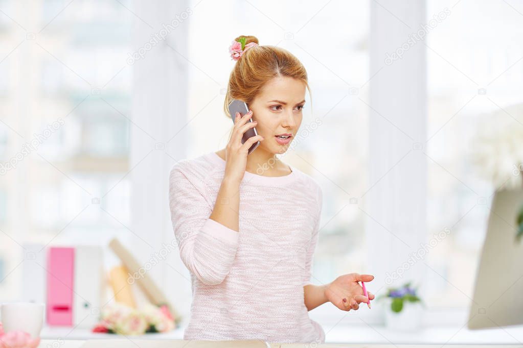 Serious interior designer with messy hair bun standing in lovely studio while talking to her colleague on smartphone