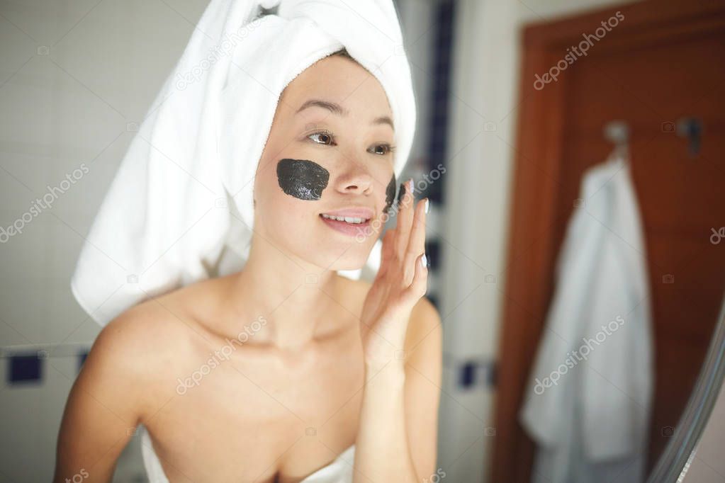 Portrait of beautiful Asian woman applying face mask during beauty treatment routine in bathroom, looking in mirror and smiling 