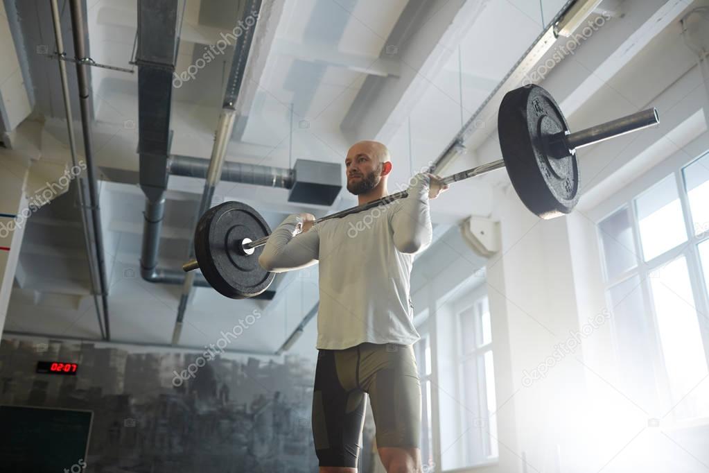 Portrait of modern strongman weightlifting with heavy barbell performing shoulder press during workout in sunlit gym