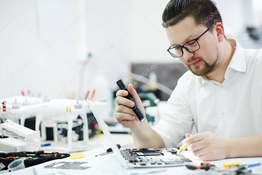 Side view portrait of man inspecting disassembled laptop with flashlight, looking for broken pats in workshop