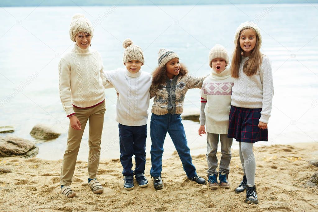 Group of cheerful children standing by lake in row posing playfully to camera on warm autumn day dressed in similar knit clothes