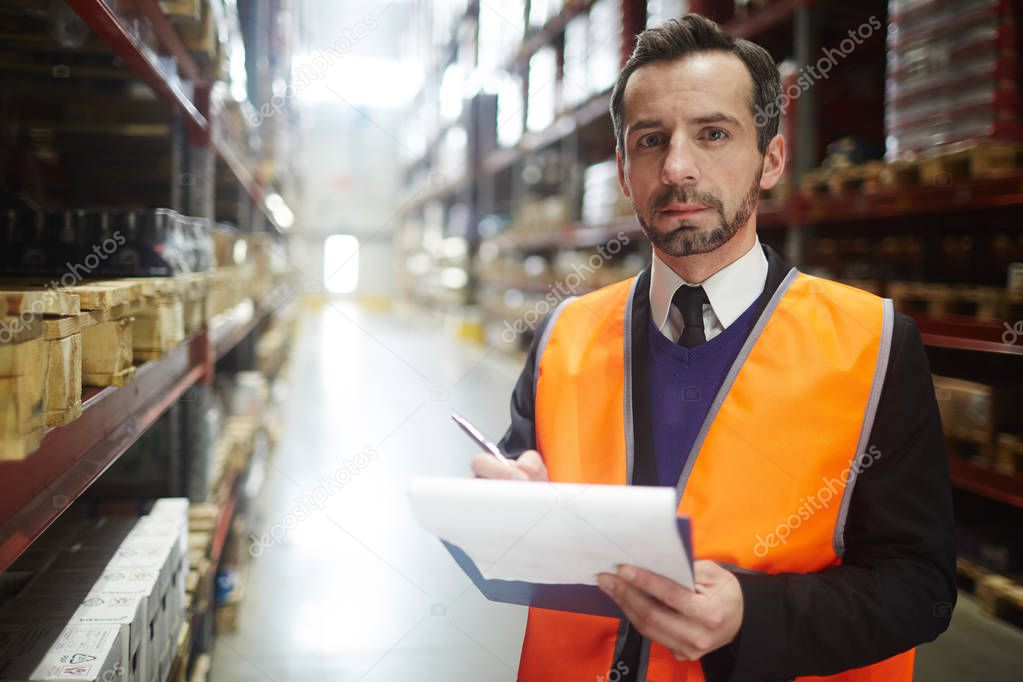 Portrait of warehouse manager looking at camera holding clipboard while doing inventory of stock