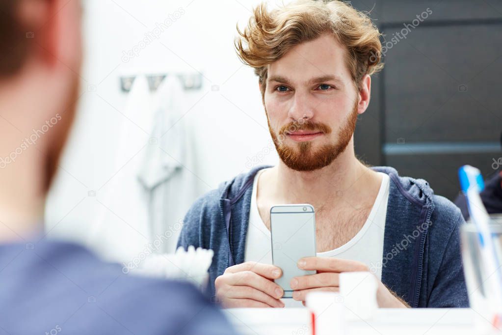 Young bearded man with smartphone looking in mirror