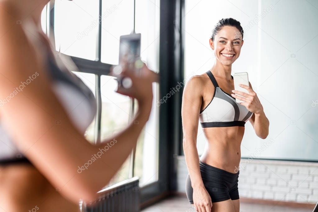 Portrait of proud muscular  woman smiling boasting her fit figure and slim waist taking selfie in gym mirror and posing