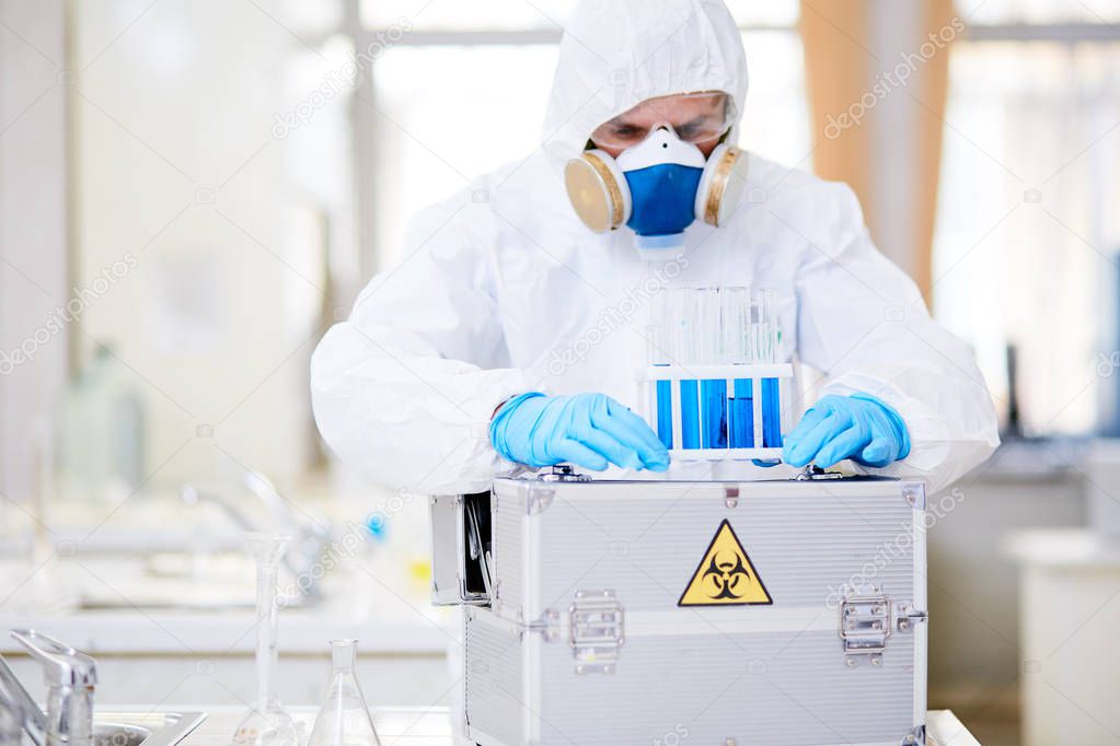 Concentrated scientist wearing hazmat suit examining test tubes while carrying out experiment at chemistry laboratory, blurred background