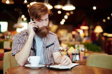 Kızıl saçlı işadamı kafeterya masada oturan ve smartphone, bel, dikey müvekkilinin için konuşurken not alma ekose desenli gömlek