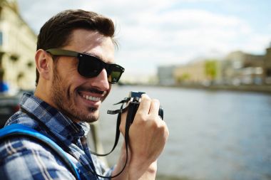 Turistik gezi tatil zevk nehir kıyısında fotoğraf çekerken kameraya bakarak gülümseyen yakışıklı genç adam giyen güneş gözlüğü portresi