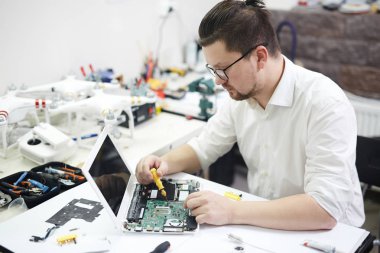 Modern adam gözlüklü elektronik ile meşgul atölyede portresi: dizüstü sökülmesi ve farklı araçları ve bölümleri tablo ile yeniden yapılandırma