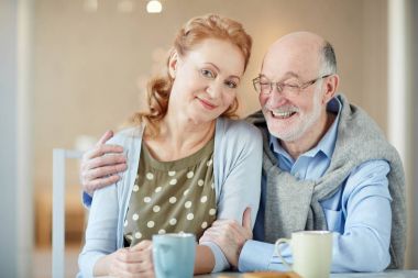 Mutlu bir evlilik: portre gülümseyen kıdemli çiftin birbirine yakın oturan, çay ve kurabiye ile mutfak masasında kucaklayan, kameraya bakıyor 