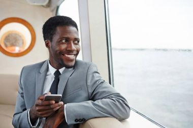 Vapur cam kenarında oturan ve içinden seyir smartphone ile mutlu Afrikalı-Amerikalı işadamı