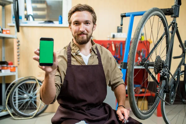微笑胡子技工的肖像坐在臀部和显示智能手机与空白屏幕摄像头 室内现代自行车修理店的背景 — 图库照片