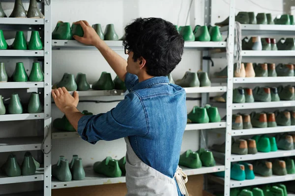 Self-employed cobbler standing in front of shelves with footwear forms and choosing model of shoes for new order