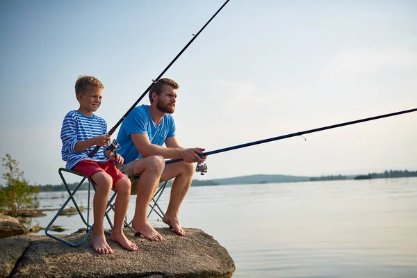 日当たりの良い夏の日釣竿とギアまだ湖の岩に座って息子と釣りにハンサムなひげを生やした父の肖像 — ストック写真