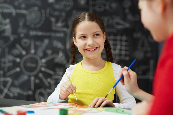 かわいい笑みを浮かべて小さな少女絵を描く黒板に対して座って学校で美術の授業中に友人との肖像画 — ストック写真