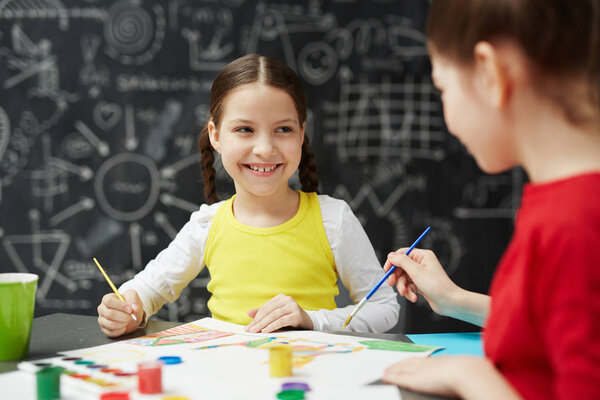 Portrait of cute smiling little girl painting pictures with friend in art studio class sitting against blackboard