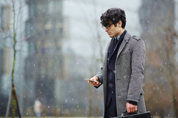 年轻的中东商人站在户外下雪和使用智能手机 看屏幕的侧面图 — 图库照片
