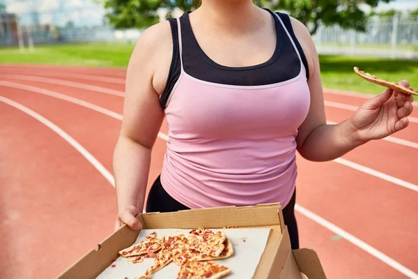 スタジアムでトレーニング中にスライスでピザを食べる黒とピンクのタンクトップのぽっちゃり女性 — ストック写真