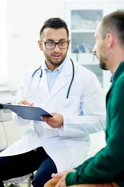 Jovem Clínico Fazendo Anotações Cartão Médico Paciente Enquanto Conversava Com — Fotografia de Stock