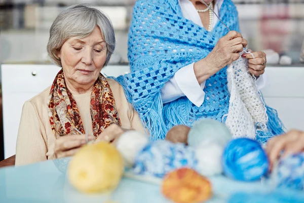集中的老年妇女坐在舒适的扶手椅和针织温暖的围巾 她的孙女 她的朋友与矢车菊蓝色披肩坐在扶手上 — 图库照片