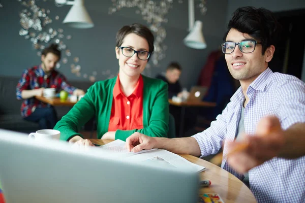 两个年轻的同事的肖像男人和女人都穿着便服和眼镜 讨论在咖啡馆开会的工作 并指向笔记本电脑屏幕 — 图库照片