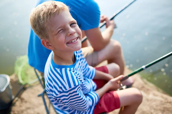欢快的小男孩的肖像 穿着条纹衬衫 眯着眼在太阳和微笑看着相机 而与父亲在湖上捕鱼 — 图库照片