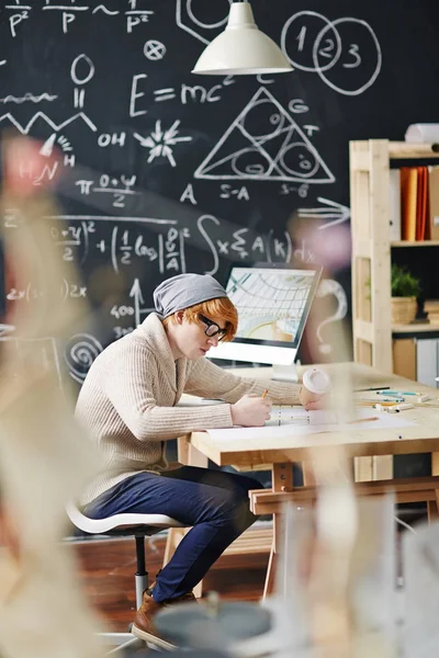 ビーニー帽子とメガネ忙しい黒板に対して大学の授業で机にチョークで書かれた科学の数式とスキームを描画を身に着けている赤髪青年像側ビュー — ストック写真
