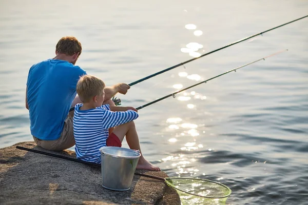 后视图的成人男子和十几岁的男孩坐在一起钓鱼与棒在晴朗的夏日湛蓝的湖面上平静的湖水中 — 图库照片