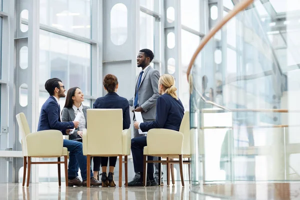集团商务人士开会时坐在圈子里 非洲人站在现代办公大楼玻璃大厅演讲 — 图库照片