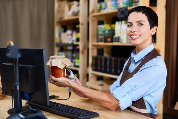 Middle-aged shop assistant looking at camera with charming smile while scanning barcode of jar with peach jam