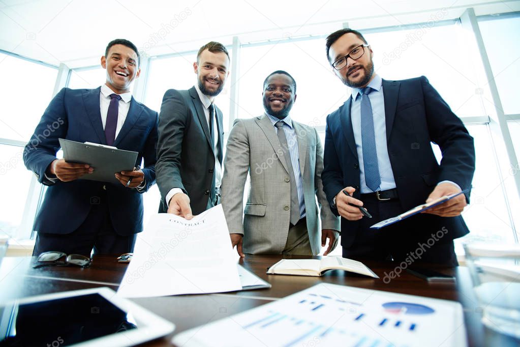 Low angle view of smiling middle-aged businessmen looking at camera while standing against panoramic window of meeting room, one of them holding contract in hand