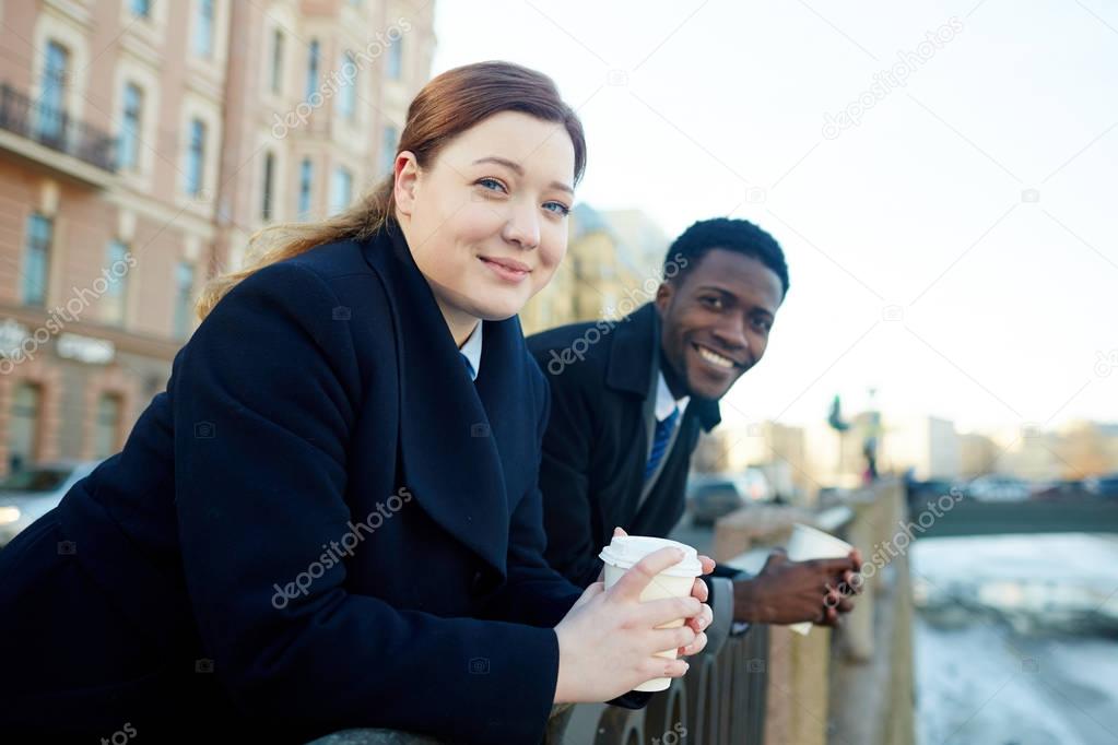 Portrait of two business colleagues in street of city, African American man and young woman,   leaning on river bank railing and looking at camera, smiling while holding disposable coffee cups both wearing coats 